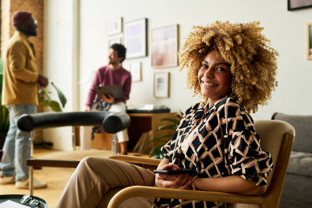 Młoda, odnosząca sukcesy Afroamerykanka dyrektor wykonawcza w eleganckich ubraniach casualwear, siedząca w fotelu i patrząca z uśmiechem na kamerę - Zdjęcie, obraz