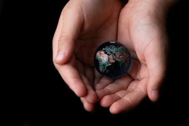 Glass symbolic globe in children's palms on a black background - 写真・画像