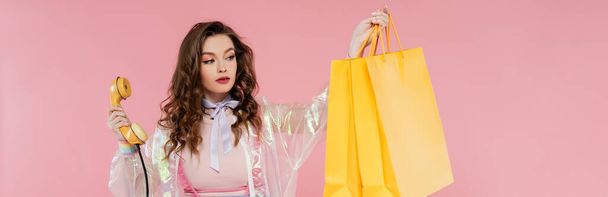 attractive woman carrying shopping bags and holding retro handset on pink background, vintage phone, concept photography, consumerism, young model in transparent jacket, banner  - Foto, Imagem