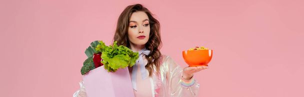 housewife concept, attractive young woman carrying grocery bag with vegetables and bowl with corn flakes, model with wavy hair on pink background, conceptual photography, home duties, banner - Фото, изображение