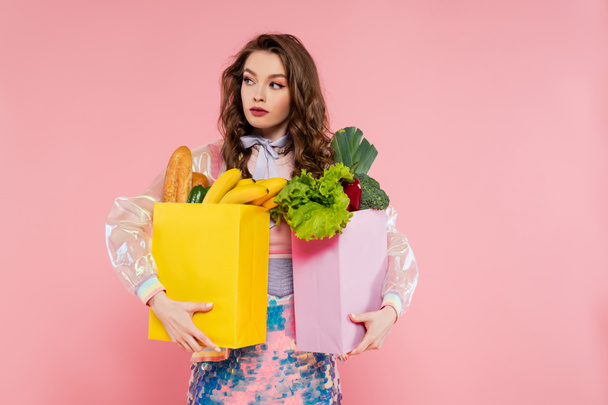 housewife concept, attractive young woman carrying grocery bags with vegetables and bananas, model with wavy hair on pink background, conceptual photography, home duties, stylish wife  - Fotoğraf, Görsel