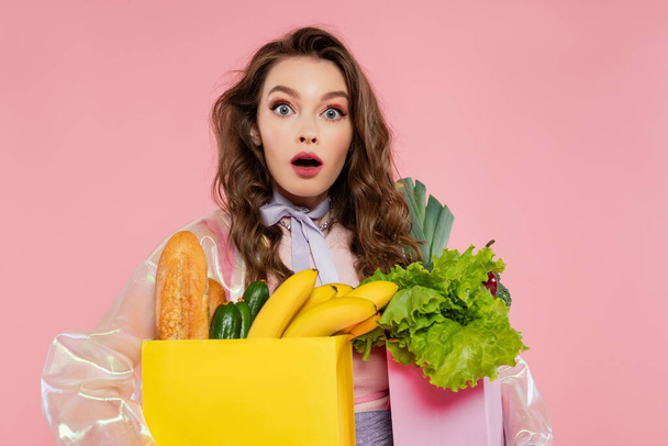 housewife concept, shocked young woman carrying grocery bags with vegetables and bananas, model with wavy hair on pink background, conceptual photography, home duties, stylish wife  - Photo, Image