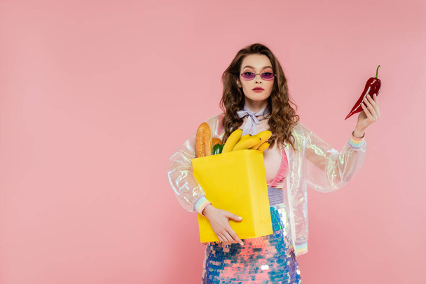 koncepcja gospodyni domowa, atrakcyjna młoda kobieta w okularach przeciwsłonecznych niosąca papierową torbę z zakupami i trzymająca czerwoną paprykę, pozująca jak lalka na różowym tle, fotografia konceptualna, obowiązki domowe - Zdjęcie, obraz