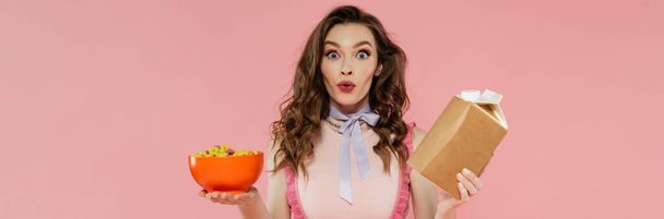 Hausfrauenkonzept, schockierte junge Frau posiert wie eine Puppe, hält Essen zum Mitnehmen und eine Schüssel mit Cornflakes, rosa Hintergrund, konzeptionelle Fotografie, häusliche Pflichten, emotional, Banner  - Foto, Bild