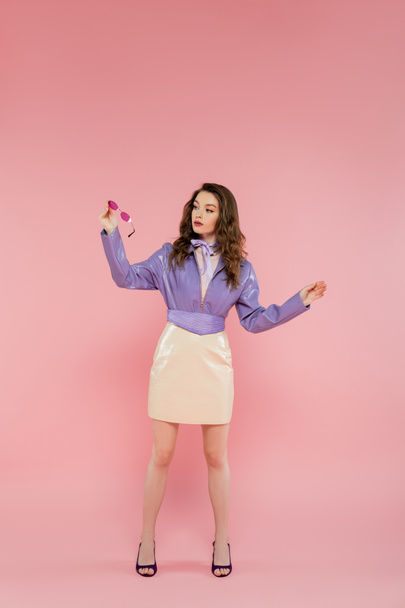 glamour, beautiful young woman gesturing and looking at sunglasses, acting like a doll, fashionable outfit, model in purple jacket and skirt, pink background, studio shot, conceptual, full length  - Фото, зображення