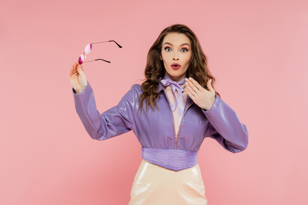glamour, surprised young woman gesturing and looking at camera, holding sunglasses, fashionable outfit, model in purple jacket and skirt standing on pink background, studio shot, acting like a doll  - Photo, Image