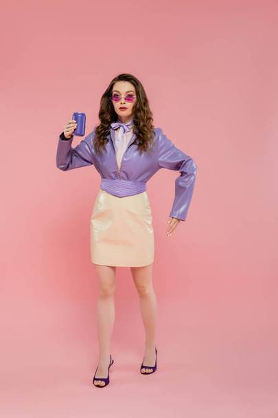 doll concept, beautiful young woman in sunglasses holding soda can and gesturing, trendy outfit, brunette model in purple jacket posing on pink background, studio shot, carbonated drink, full length  - Foto, Imagem