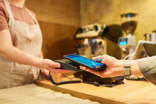 Close-up of clerk with terminal over counter desk and female client with smartphone during contactless payment for purchase in cafeteria - Φωτογραφία, εικόνα
