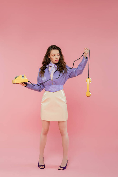 doll pose, beautiful young woman with wavy hair looking at handset while holding yellow retro phone, brunette model in purple leather jacket posing on pink background, studio shot, full length - 写真・画像