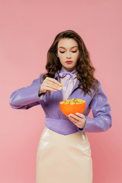 concepto de fotografía, mujer morena con el pelo ondulado fingiendo ser una muñeca, sosteniendo tazón con hojuelas de maíz, mirando el desayuno, posando sobre fondo rosa, chaqueta púrpura de moda - Foto, Imagen