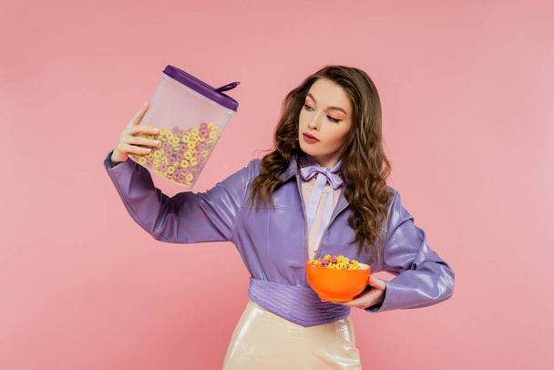 concepto de fotografía, mujer morena con el pelo ondulado fingiendo ser una muñeca, sosteniendo contenedor con hojuelas de maíz, sabroso desayuno, posando sobre fondo rosa, elegante chaqueta púrpura - Foto, imagen