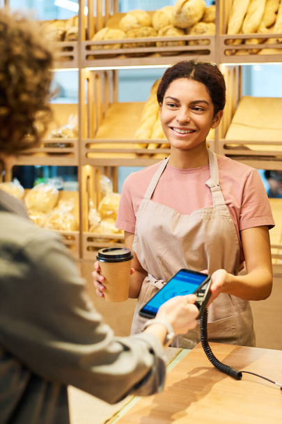 Happy young female clerk with cup of coffee looking at client of bakery shop with smile during process of contactless payment for purchase - 写真・画像