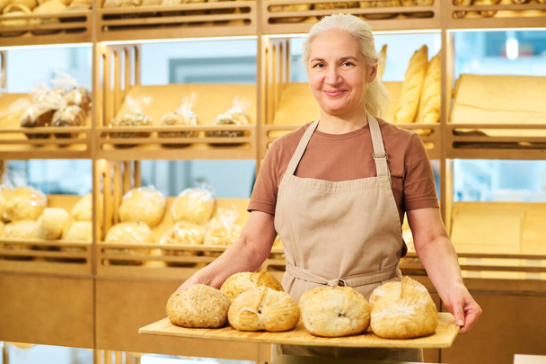 Щаслива зріла жінка-пекарня або клерк в фартусі тримає дерев'яний лоток зі свіжим запеченим пшеничним хлібом і дивиться на камеру проти дисплея - Фото, зображення