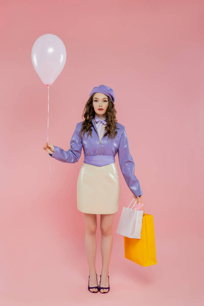 moda fotografia, atrakcyjne i młoda kobieta trzyma balon i torby na zakupy na różowym tle, pozowanie jak lalka, stojąc i patrząc w aparat, modne, konsumpcjonizm, fioletowy strój  - Zdjęcie, obraz
