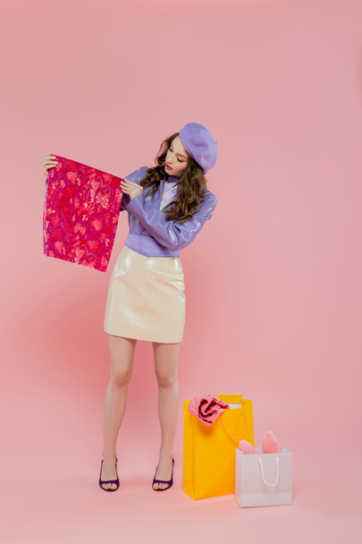 fashion and consumerism, attractive young woman in beret holding vibrant color clothes near shopping bags on pink background, consumerism, standing in trendy leather jacket and skirt, full length - Foto, Bild