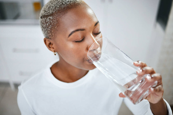 Gezondheid, glas en vrouw drinkwater voor hydratatie, wellness en vloeibare voeding bij haar thuis. Dorst, energie en Afrikaanse vrouwelijke persoon genieten van koude gezonde drank in de keuken van haar appartement - Foto, afbeelding