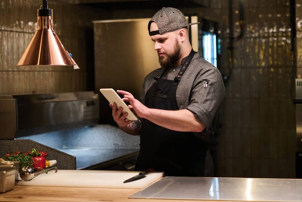 Młody poważny kucharz w mundurze za pomocą tabletu szukać receptur online pizzy stojąc przy miejscu pracy ze świeżych składników - Zdjęcie, obraz