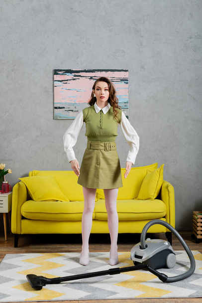 housekeeping concept, young woman with wavy hair standing on clean carpet near vacuum cleaner and yellow couch, gesturing and looking at camera, housewife in dress, domestic life, posing like a doll  - Zdjęcie, obraz