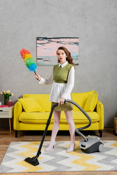 housekeeping concept, young woman with wavy hair standing on carpet and holding dust brush, using vacuum cleaner, housewife in dress and white tights, domestic life, posing like a doll  - Foto, imagen