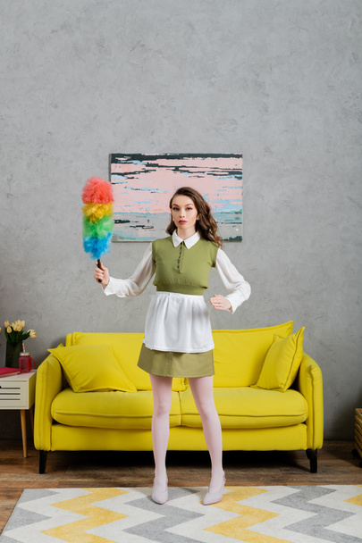 housekeeping concept, young woman with wavy hair standing and holding dust brush, housewife in dress and white tights looking at camera, gesturing unnaturally, domestic life, posing like a doll  - Foto, Bild