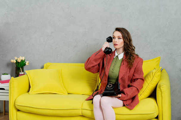 retro vibrace, atraktivní žena s vlnitými vlasy sedící na žlutém gauči, hospodyňka mluví na retro telefonu, pózuje jako panenka, dívá se jinam, moderní interiér, obývací pokoj  - Fotografie, Obrázek