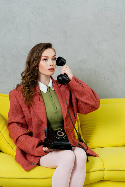 vintage vibes, beautiful woman with brunette and wavy hair sitting on yellow couch, housewife talking on retro telephone, posing like a doll, looking away, modern interior, living room  - Фото, изображение