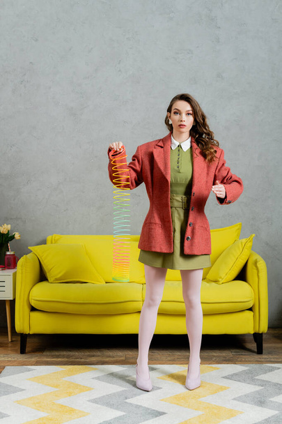 beautiful woman posing like a doll and playing with rainbow slinky, looking at camera, modern living room with yellow couch, childish, vintage, nostalgia, colorful toy, leisure and fun  - Foto, imagen