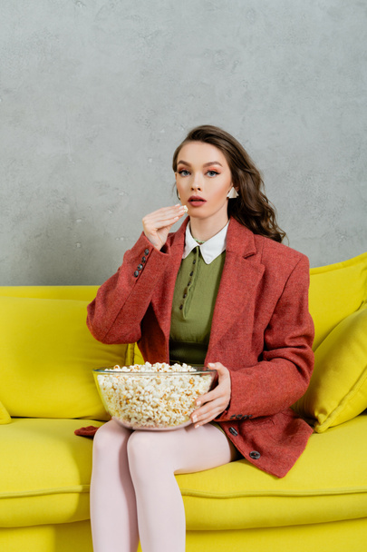 pretty girl acting like a doll, concept photography, young woman with brunette wavy hair eating popcorn, holding bowl, salty snack, home entertainment, sitting on comfortable yellow sofa  - Photo, Image