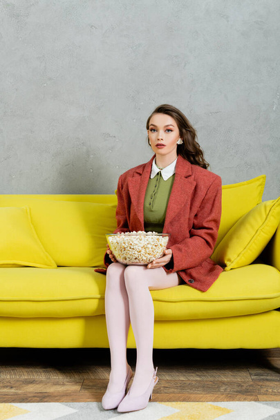 concept fotografie, jonge vrouw met brunette golvend haar holding bowl met popcorn, zoute film snack, home entertainment, zitten op comfortabele gele bank en kijken naar de camera - Foto, afbeelding