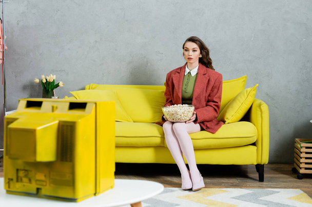 pretty girl acting like a doll, concept photography, young woman with brunette wavy hair sitting with bowl of popcorn, watching tv, home entertainment, sitting on comfortable yellow sofa  - 写真・画像