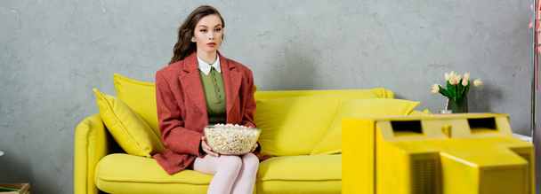 Frau, die sich wie eine Puppe benimmt, Konzeptfotografie, attraktives Mädchen mit brünetten welligen Haaren, sitzend mit einer Schüssel Popcorn, fernsehend, Home Entertainment, sitzend auf einem bequemen gelben Sofa, Banner  - Foto, Bild