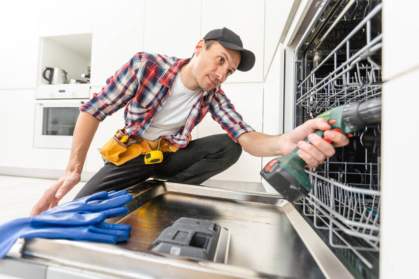 Ένας επισκευαστής διορθώνει δυσλειτουργικό πρόβλημα πλυντηρίου πιάτων κουζίνας. Υψηλής ποιότητας φωτογραφία - Φωτογραφία, εικόνα