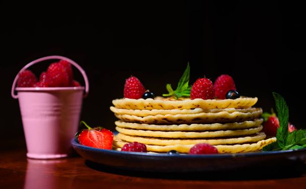 Stillleben mit einem Teller Pfannkuchen mit frischen Beeren und Minzblättern in der Nähe eines rosafarbenen Eimers mit Erdbeeren, isoliert vor dunklem Hintergrund. Fastnacht. Faschingsdienstag. Pfannkuchen-Tag. Hintergrund der Nahrungsmittel - Foto, Bild