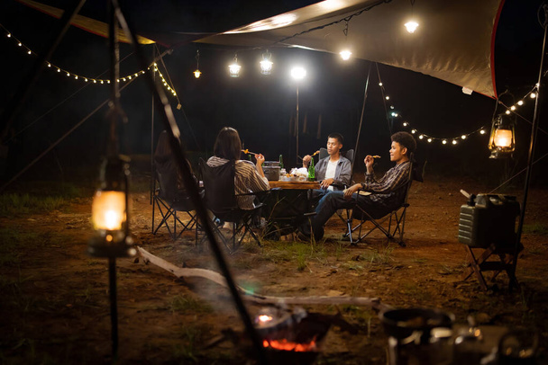 湖の休日の夜にキャンバスの下でバーベキューを食べる若いアジア人は、ライトの下にテントを持っています. - 写真・画像