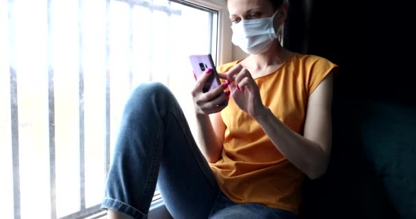 Nainen suojaava naamio hänen kasvonsa istuu ikkunalaudalla puhelin käsissään 4k elokuva. Uusi todellisuus pandemia covid19 käsite - Materiaali, video