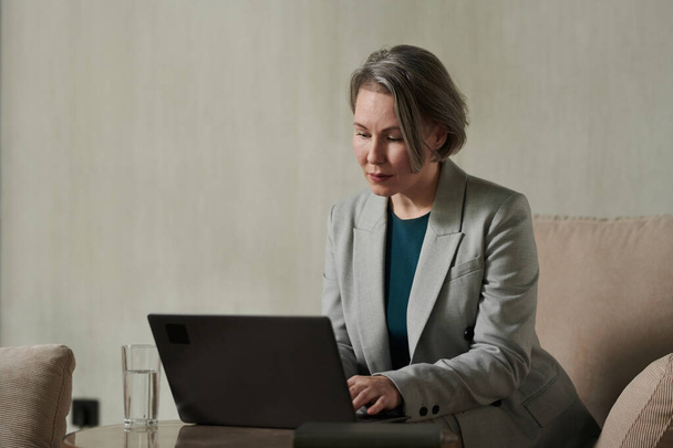 Psicólogo maduro grave em formalwear sentado em poltrona no escritório e digitando no teclado do laptop enquanto consulta pacientes on-line - Foto, Imagem