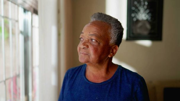 Kontemplativní starší žena stojí u okna a dívá se ven. Zamyšlený zamyšlený výraz afroamerického staršího člověka v 70. letech - Fotografie, Obrázek