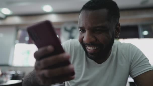 Un brasileño negro feliz sosteniendo el dispositivo celular sonriendo. Persona afroamericana mirando la pantalla del teléfono - Imágenes, Vídeo