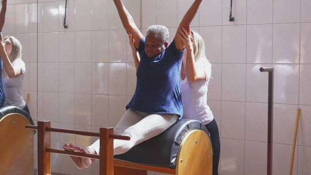 Female Pilates coach assisting an African American elderly woman to stretch body in Physiotherapy exercise session. Strength and flexibility old age workout lifestyle routine - Photo, Image