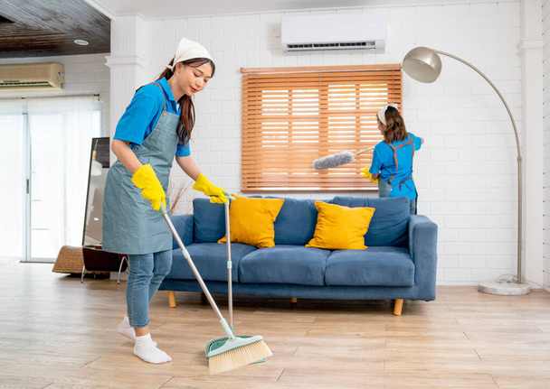 アジアの家政婦や家政婦のチームの広いショットは、 1つの使用ほうきのきれいな床や窓を掃除するための他の使用羽のダスターと一緒にリビングルームをきれいにするのに役立ちます. - 写真・画像
