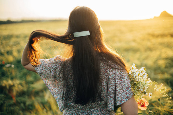 Piękna kobieta z dzikimi kwiatami ciesząca się zachodem słońca na polu jęczmienia. Atmosferyczny moment spokoju, rustykalne powolne życie. Stylowe kobiety zbierające dzikie kwiaty i relaksujące w letniej wsi - Zdjęcie, obraz