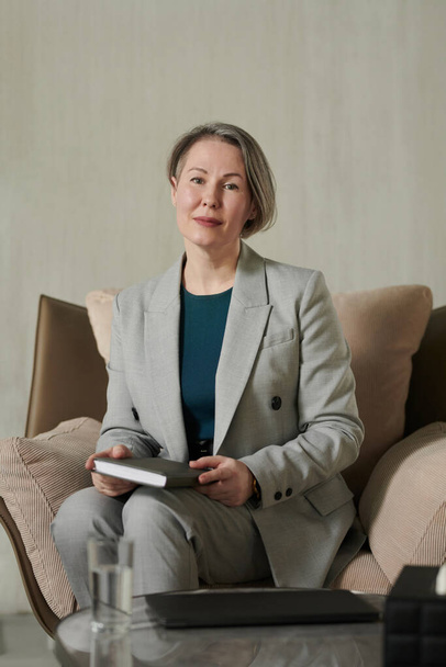 Zelfverzekerde glimlachende vrouwelijke psycholoog in grijs pak zittend in zachte comfortabele fauteuil door haar werkplek en kijkend naar camera - Foto, afbeelding