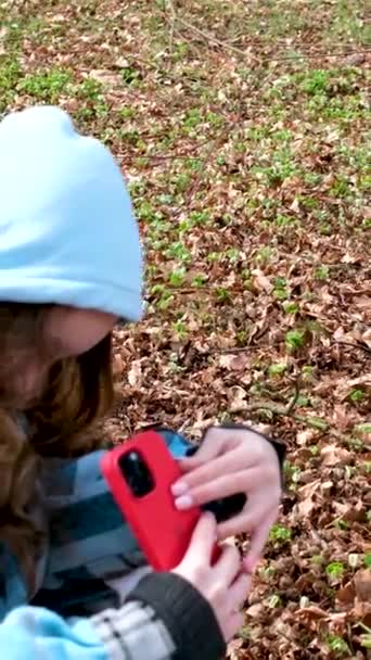 syksyllä tai keväällä laskenut lähtee tyttö punaisella puhelimella tekee kuvia alhaalta ylöspäin punainen kotelo matkapuhelimeen hupullinen ruudullinen sininen takki teini puistossa - Materiaali, video