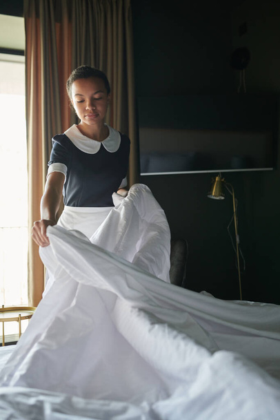 Young chambermaid in uniform standing by double bed in hotel room and changing blanket and other bedclothes before arrival of new guests - 写真・画像
