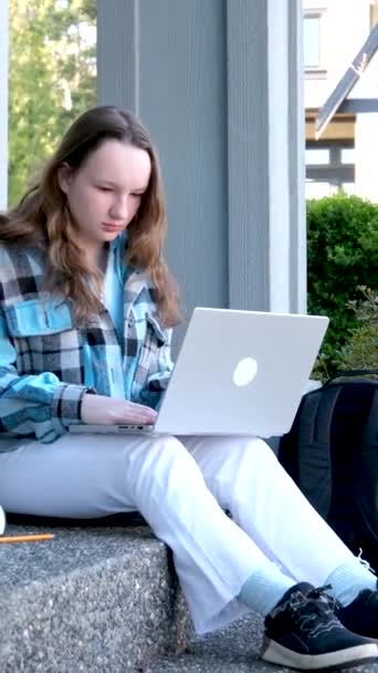 Sit alone on the verch of the house slow motion video white jeans black tenisky laptop in hands autumn spring jacket checkered blue loose hair private sector teenager use laptop next to folder - Záběry, video