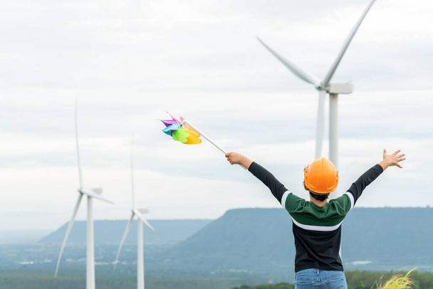 Progressive young asian boy playing with wind pinwheel toy in the wind turbine farm, green field over the hill. Green energy from renewable electric wind generator. Windmill in the countryside concept - Photo, Image