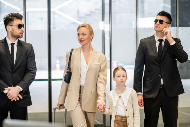 personal security concept, two bodyguards in suits and sunglasses protecting happy blonde woman and child, luxury lifestyle, woman and girl holding hands and walking inside of hotel  - Photo, Image