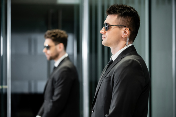 bodyguard service, private security, professional in suit and sunglasses standing in hotel lobby near work partner, earpiece, communication, luxury hotel, vigilance, protection and work, side view - Foto, Imagem
