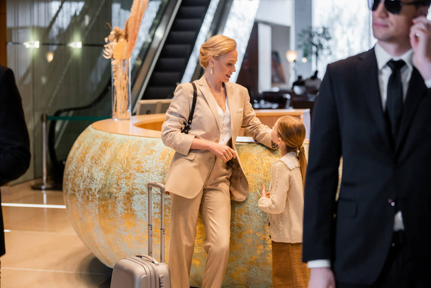 personal security concept, blonde woman standing at reception desk with preteen daughter, rich lifestyle, family travel, bodyguard in suit and sunglasses standing on blurred foreground, safety - Fotó, kép