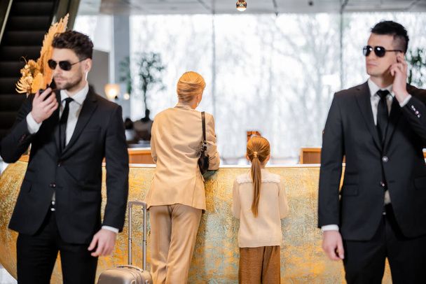 personal security, lifestyle, blonde mother with child standing at reception desk, woman and preteen girl during check in, bodyguards in suit and sunglasses protecting rich family in hotel  - Photo, Image